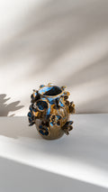 A handmade ceramic vase with a textured, organic shape, decorated with a glossy blue and gold floral glaze, set against a white wall with soft leaf shadows.