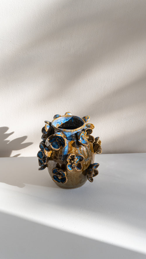 A handmade ceramic vase with a textured, organic shape, decorated with a glossy blue and gold floral glaze, set against a white wall with soft leaf shadows.
