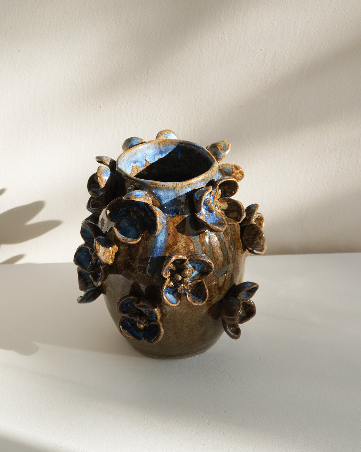 A small handmade ceramic vase with a dark brown and black textured glaze, decorated with raised floral or leaf-like protrusions, set against a warm off-white wall with soft diagonal shadows.