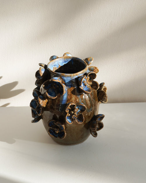 A small handmade ceramic vase with a dark brown and black textured glaze, decorated with raised floral or leaf-like protrusions, set against a warm off-white wall with soft diagonal shadows.