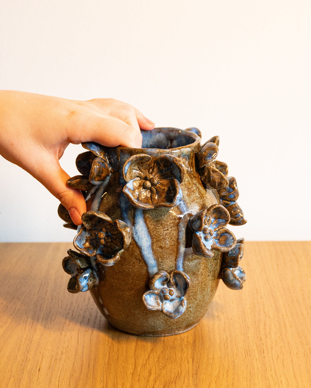 A hand holding a small ceramic vase with a brown and blue floral glaze, set on a wooden table against a plain light background.