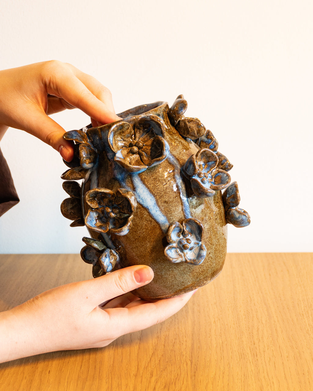 A handmade ceramic pot with a textured, organic floral design and glossy dark brown glaze, held by hands over a wooden table against a plain white wall.
