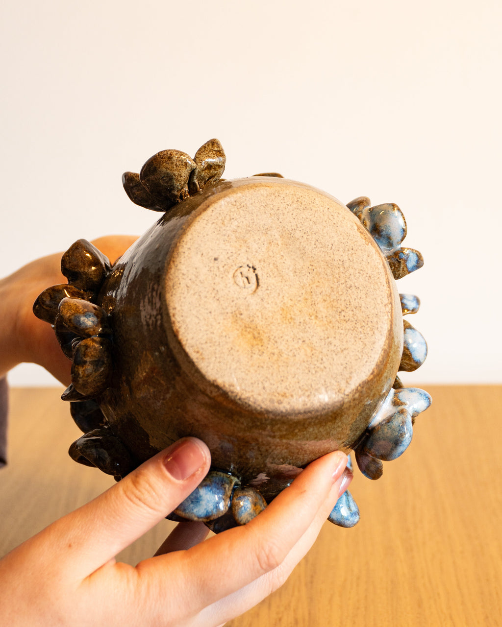 A handmade ceramic pot with a brown and blue glazed finish, held by two hands over a wooden table against a plain beige wall.