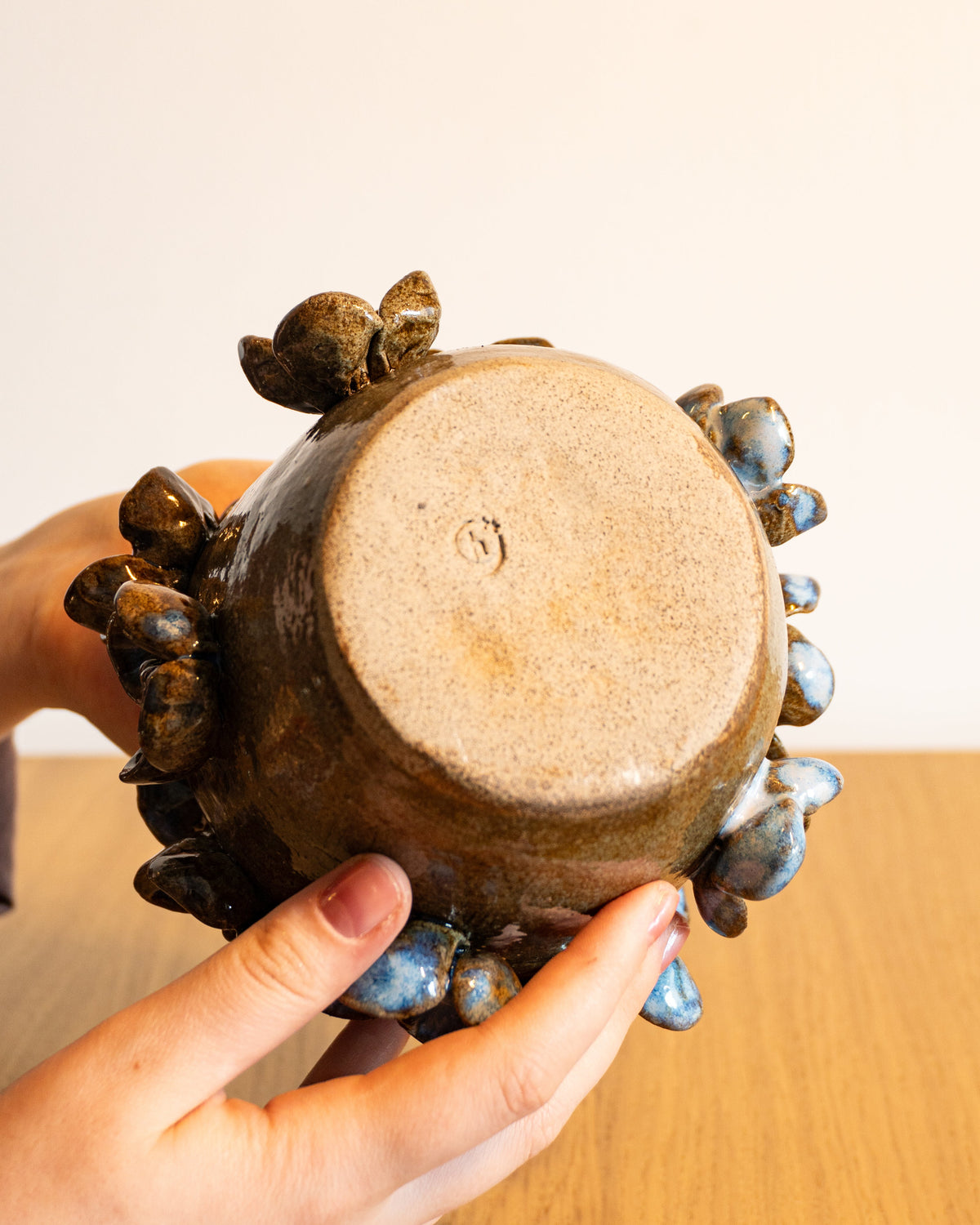 A handmade ceramic pot with a brown and blue glazed finish, held by two hands over a wooden table against a plain beige wall.