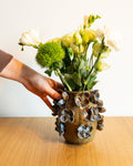 A hand holds a rustic brown ceramic vase decorated with blue and white floral details, filled with white and green flowers, set on a light wooden table against a plain white wall.