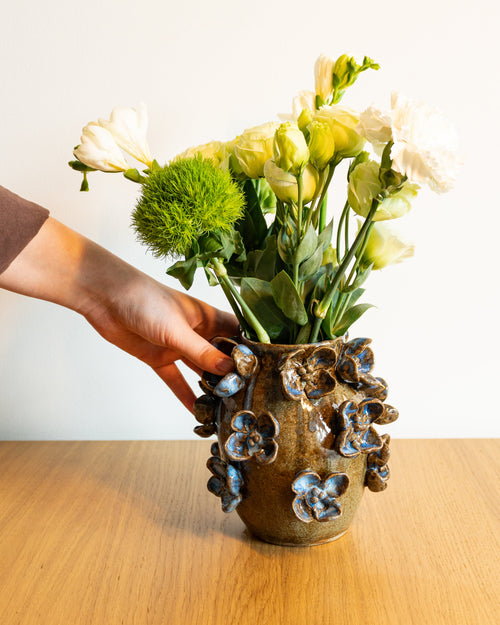 A hand holds a rustic brown ceramic vase decorated with blue and white floral details, filled with white and green flowers, set on a light wooden table against a plain white wall.