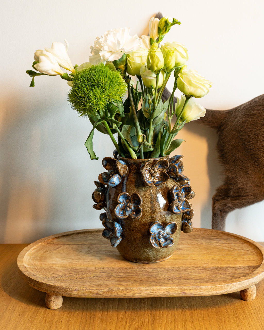 A rustic brown ceramic flower vase with blue glazed floral details, filled with white and pale yellow flowers, set on a round wooden tray against a plain light wall.