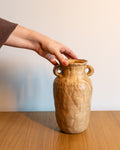A hand holding a small rustic ceramic vase with two loop handles, set on a wooden table against a plain light wall.