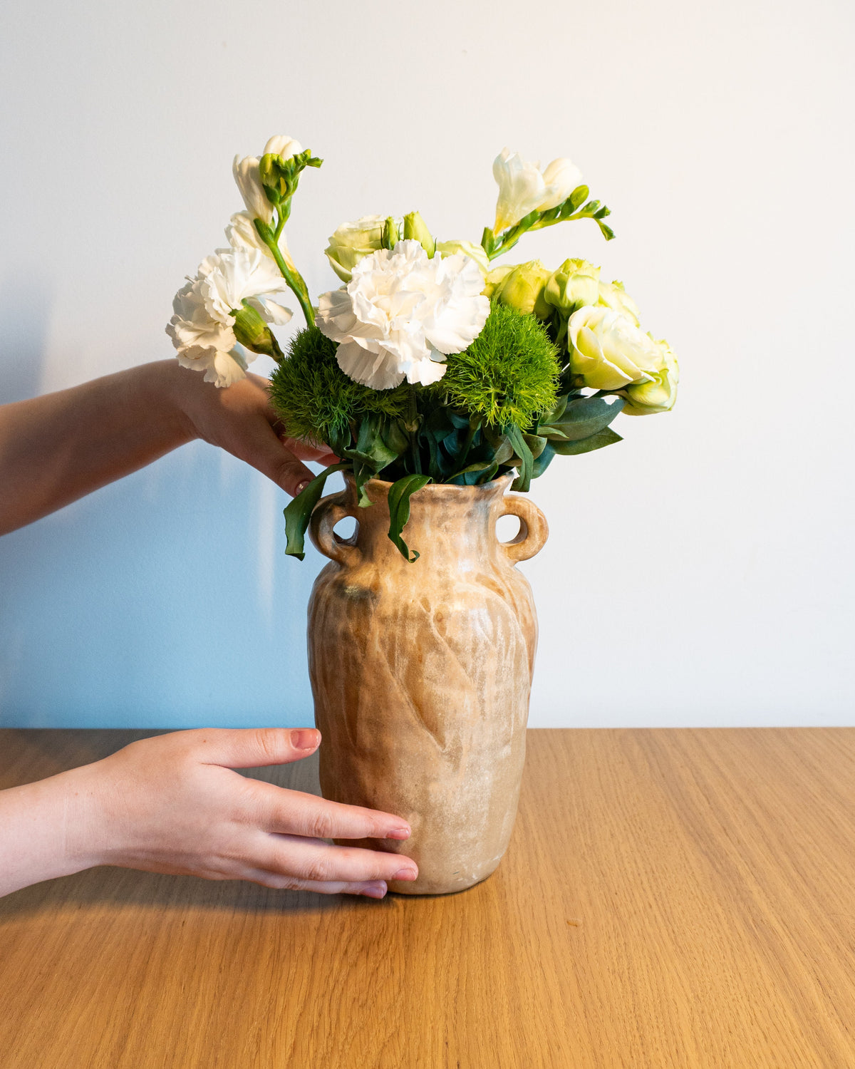 A rustic beige ceramic vase with two loop handles, filled with white flowers and green foliage, sitting on a wooden table with hands adjusting it against a plain light blue wall.