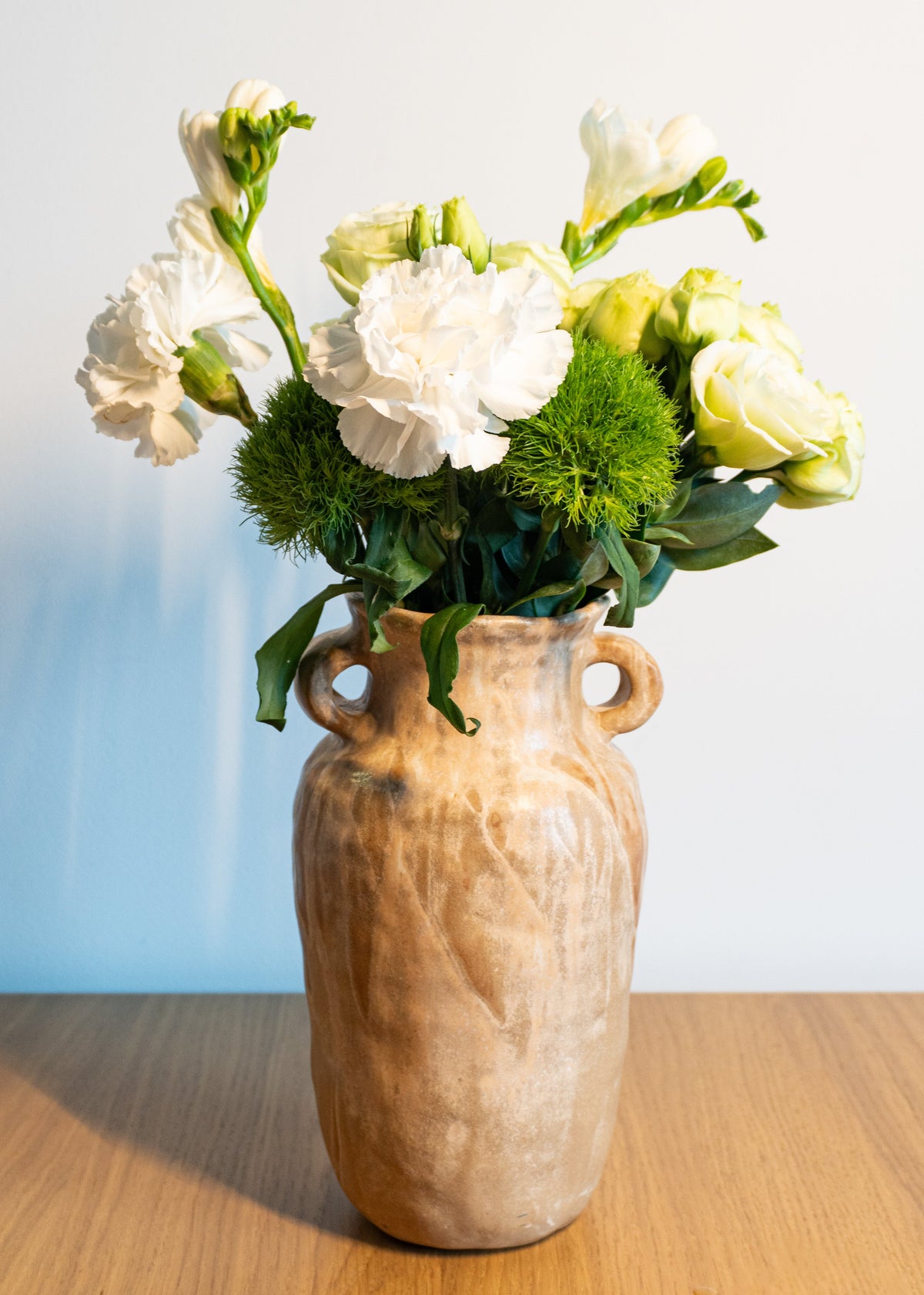 A rustic beige ceramic vase with two side handles, filled with a bouquet of white carnations, pale yellow roses, and green thistle stems, set on a wooden tabletop against a light blue and white background.