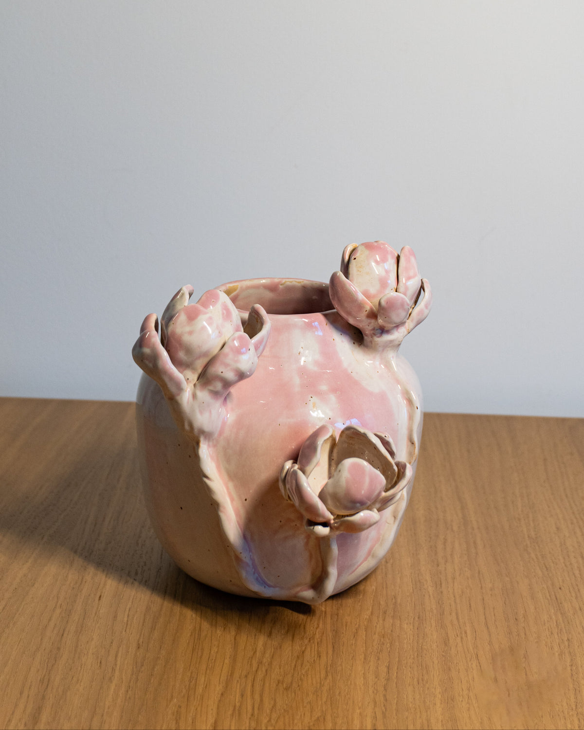 A handmade ceramic pot with a pink and beige glaze, featuring three sculpted flower buds rising from the top, displayed on a wooden tabletop against a plain light gray wall.