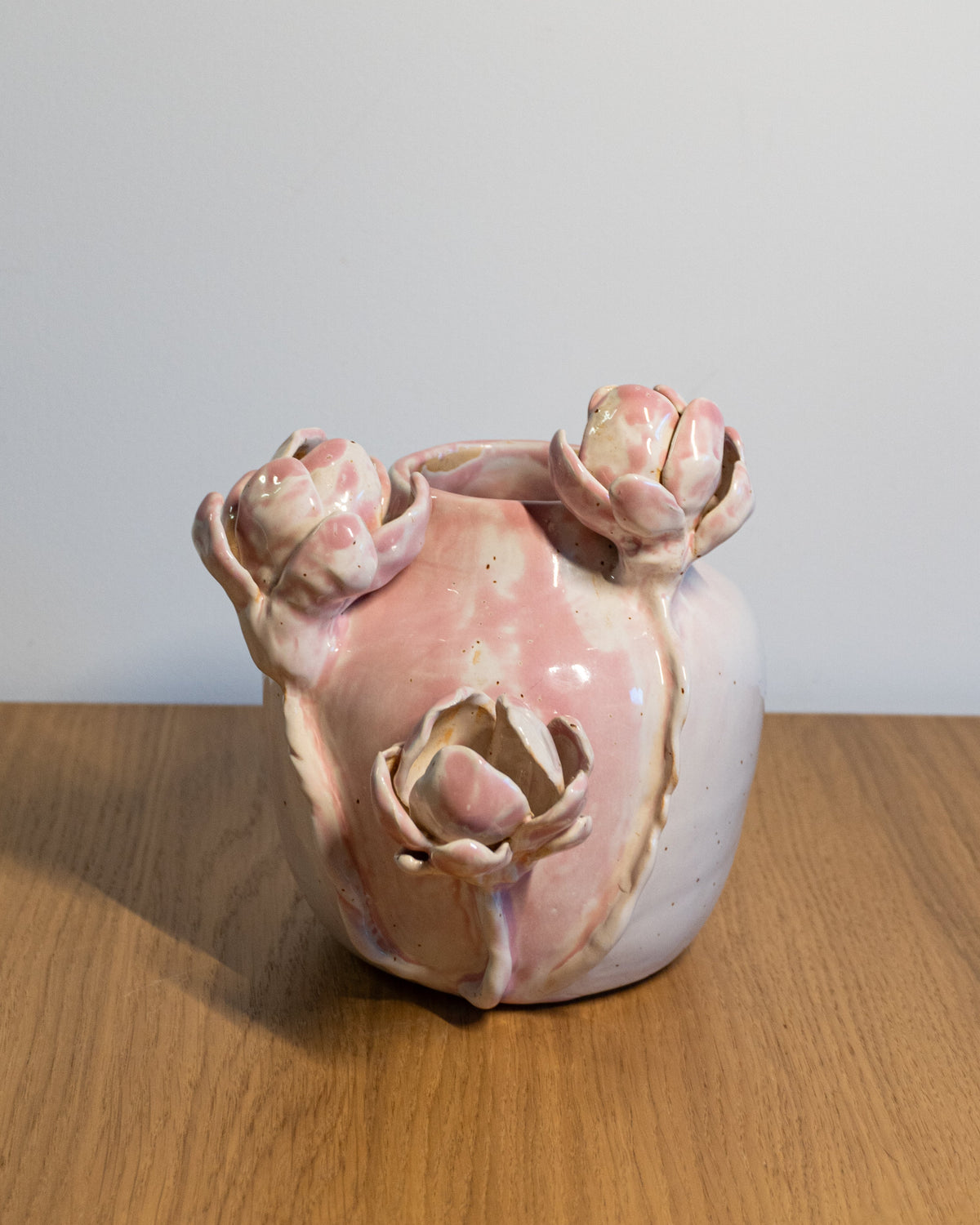 A handmade ceramic vase with a pink and white glaze, featuring sculpted rose buds on the rim and a central floral relief, displayed on a wooden surface against a plain light gray wall.