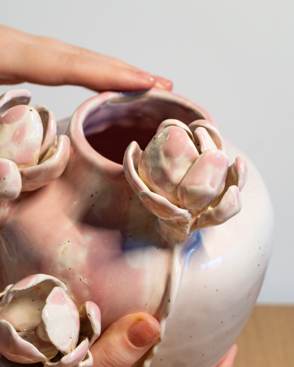 A close-up of a handmade ceramic vase with a rounded body and three protruding flower-like sculptural details, being held by hands against a neutral light gray background.
