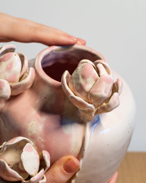 A close-up of a handmade ceramic vase with a rounded body and three protruding flower-like sculptural details, being held by hands against a neutral light gray background.