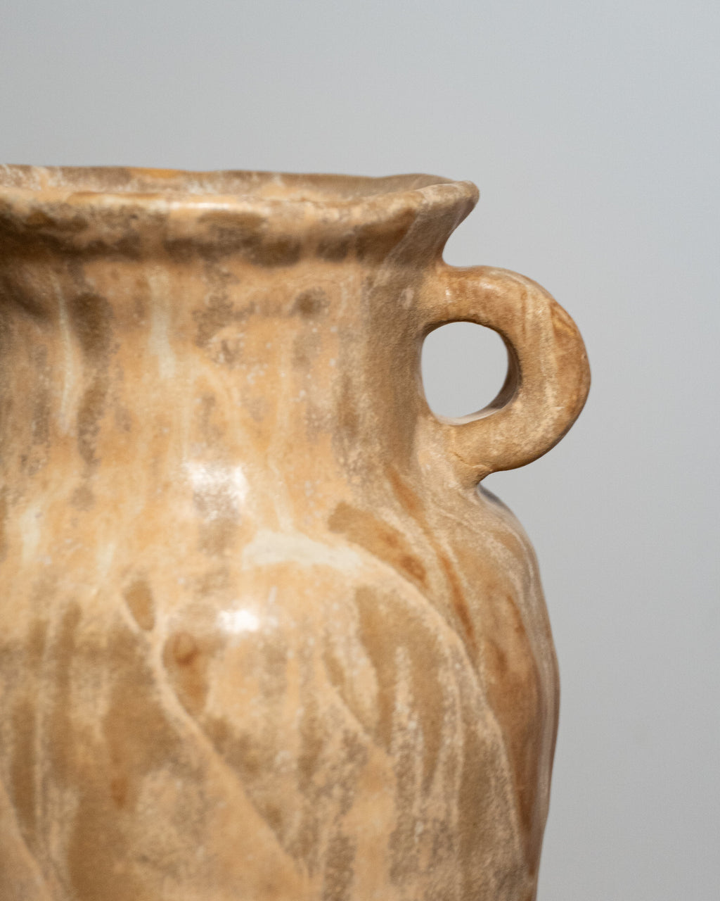 Close-up of a ceramic vase with a single loop handle, featuring a textured, organic marbled pattern in warm beige, tan, and cream tones against a plain light gray background.