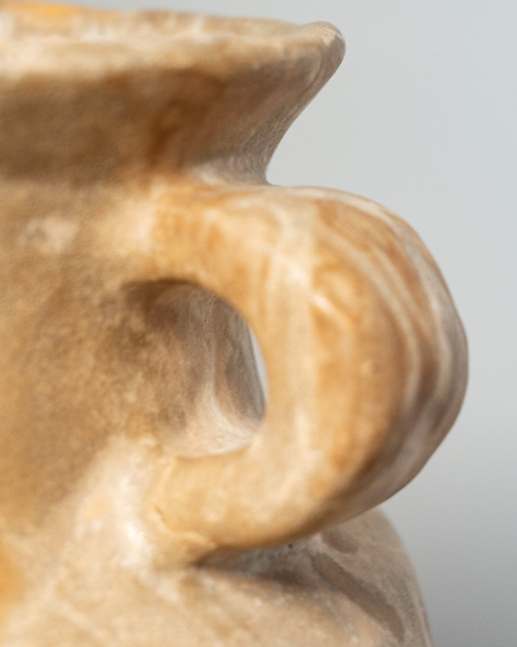 Close-up detail of a beige stone or ceramic vessel with a rounded handle and smooth matte finish against a soft gray background.