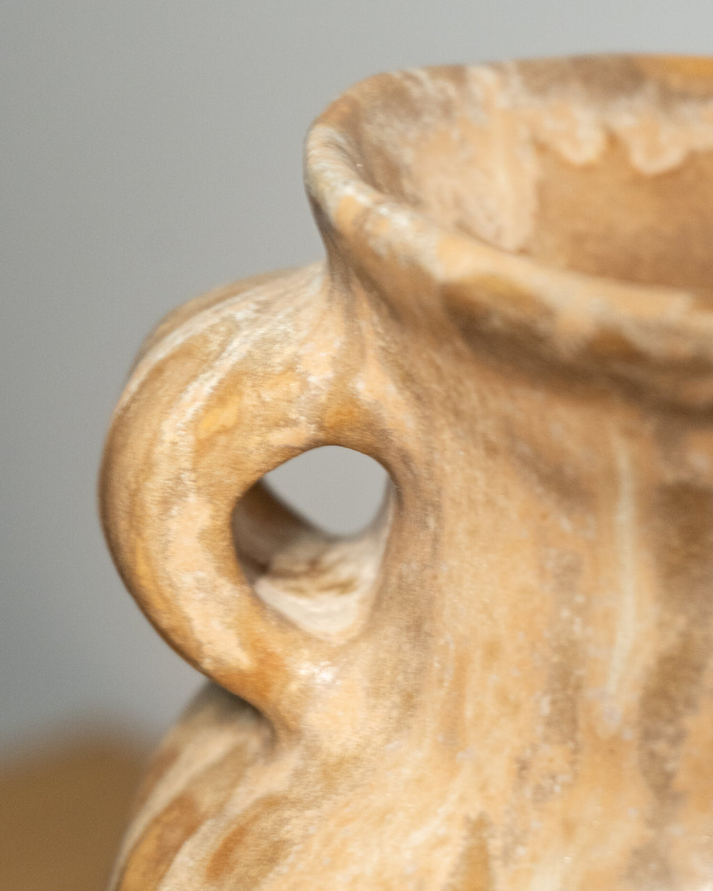 Close-up of a textured ceramic vase in a warm sandy beige color, showing the side handle and part of the open rim against a soft gray background.