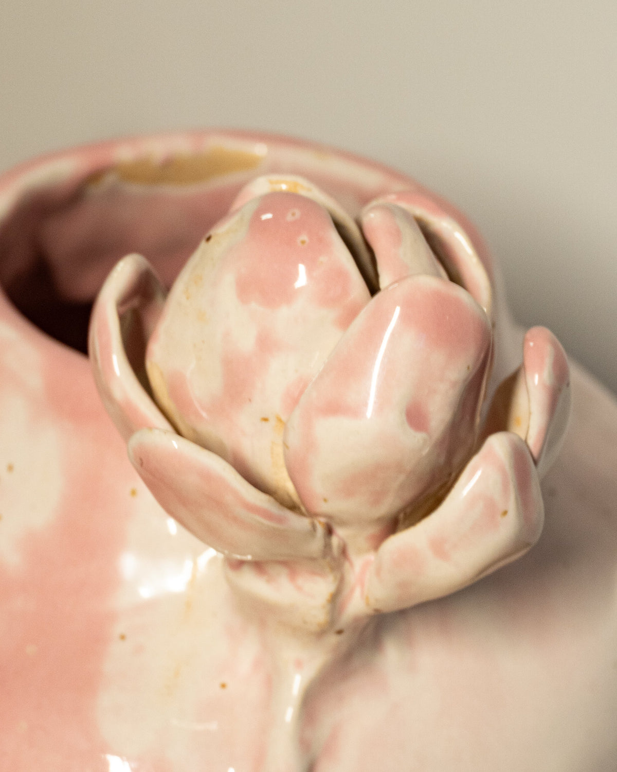 Close-up of a glossy pink ceramic vase with a sculpted flower on top, showing a soft blush and white marbled glaze against a neutral background.
