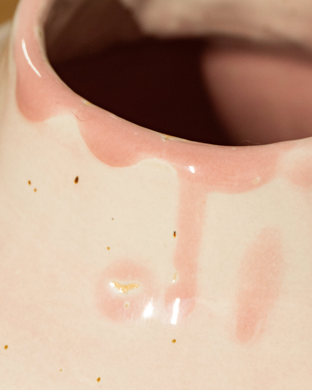Close-up of a glossy ceramic vase with a pale pink and cream glaze, showing dripping pink glaze around the rim and a few small brown specks on the surface.