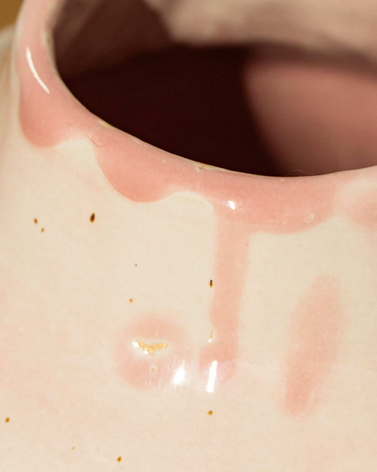 Close-up of a glossy ceramic vase with a pale pink and cream glaze, showing dripping pink glaze around the rim and a few small brown specks on the surface.