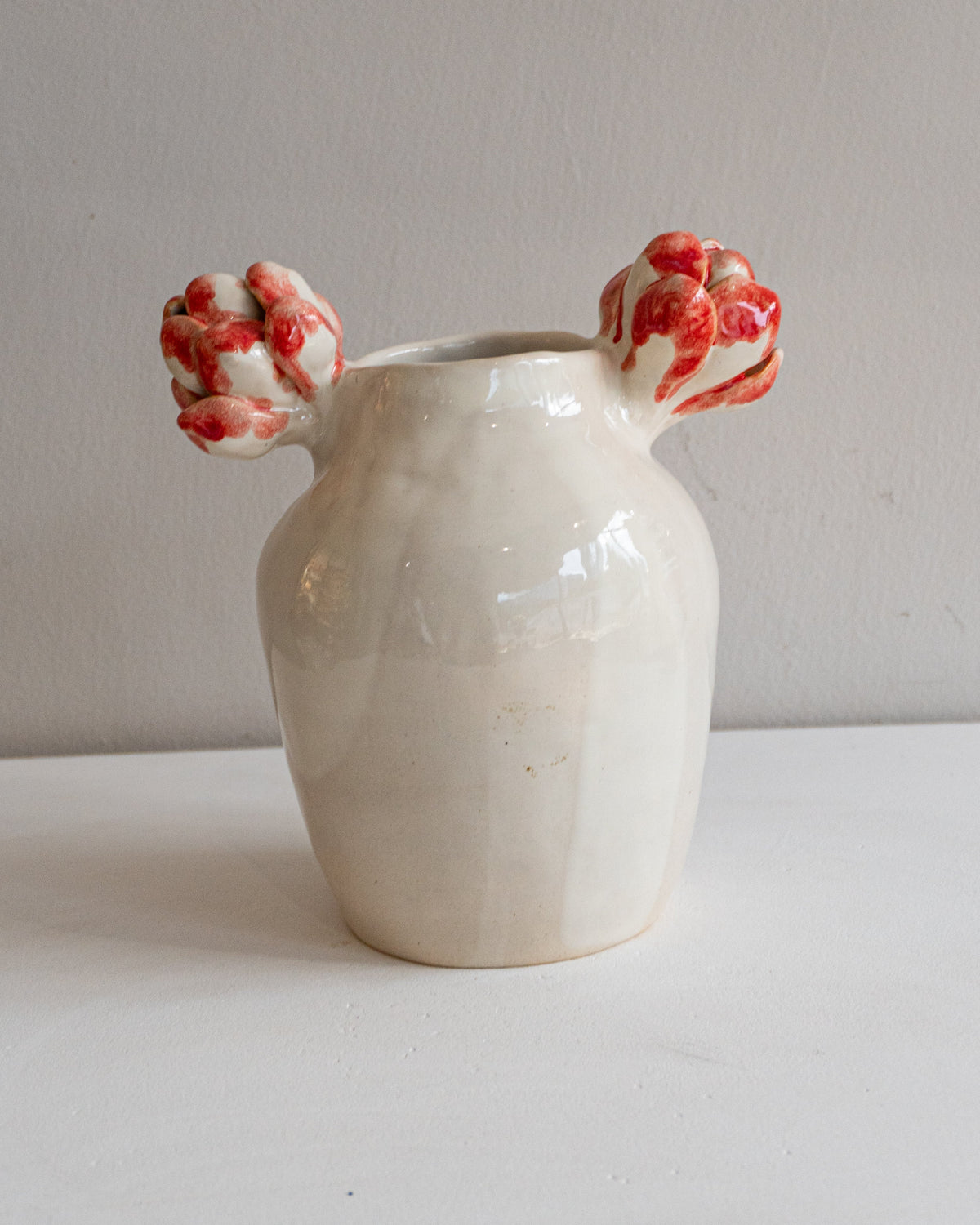 A small handmade ceramic vase with a matte off-white body and two red-and-white patterned floral handles, set against a plain light gray wall and white surface.