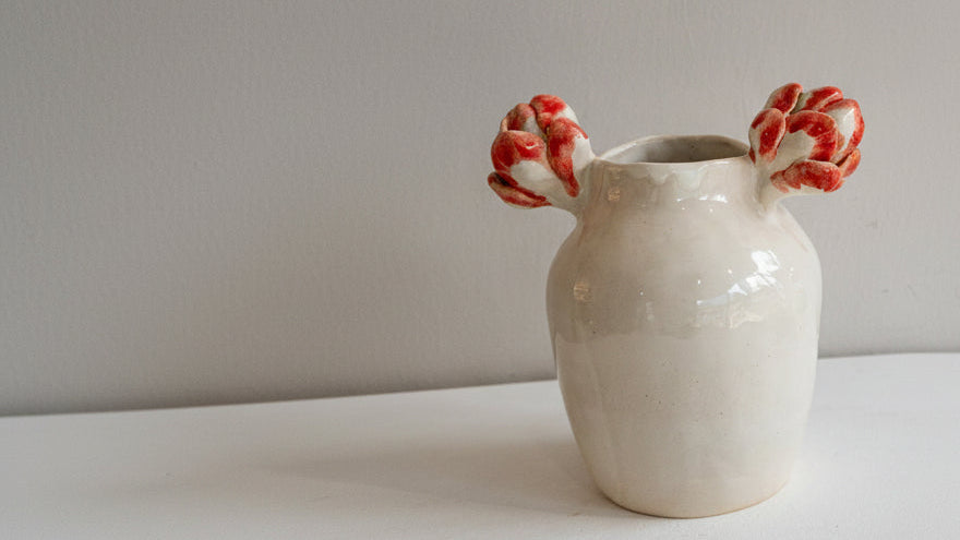 A handmade ceramic vase with a glossy off-white body and two sculpted red and white flower handles, set against a neutral light gray background.