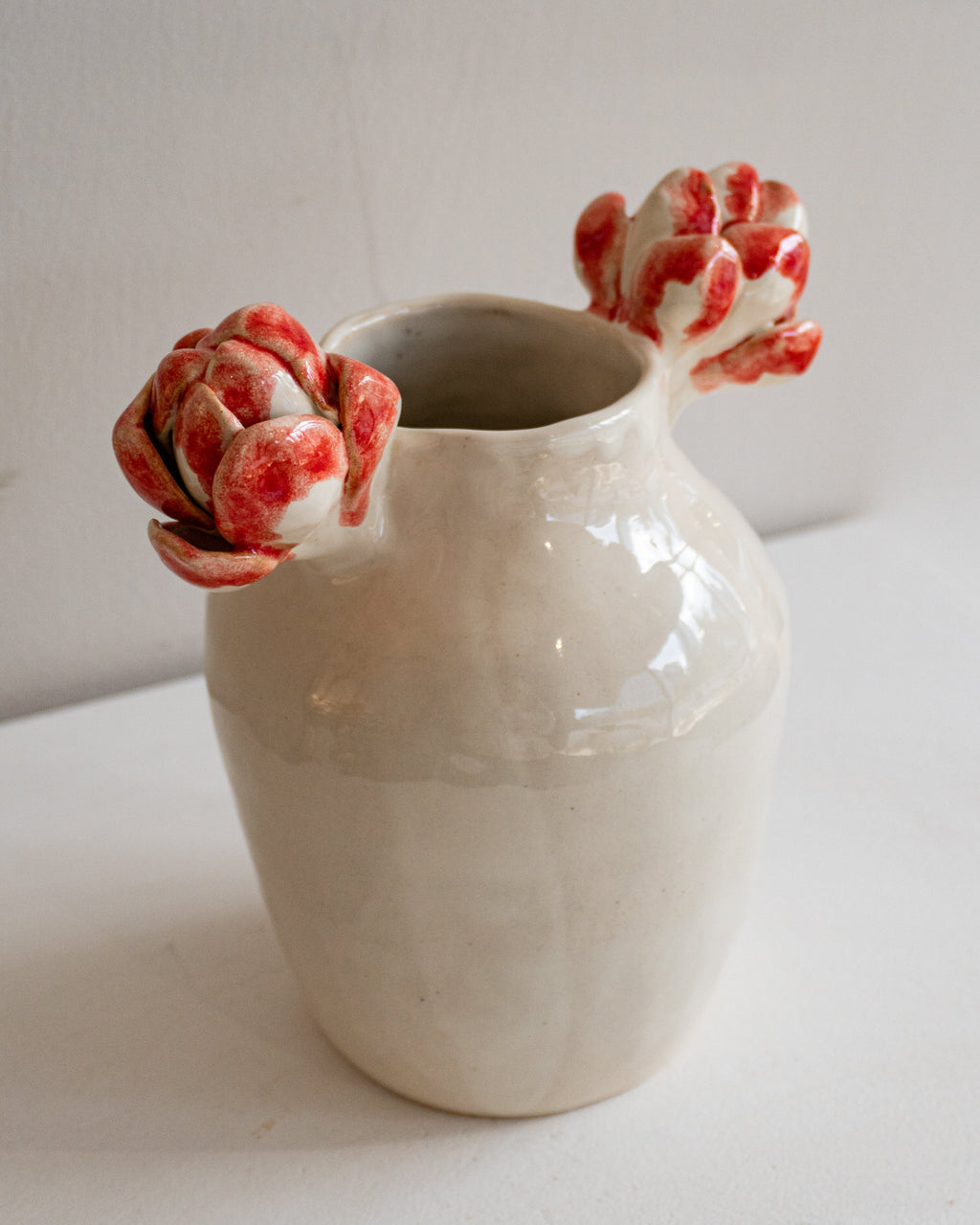 A small glossy ceramic vase in warm off-white with two sculptural red-and-white flower-like protrusions at the rim, photographed against a plain light wall and surface.