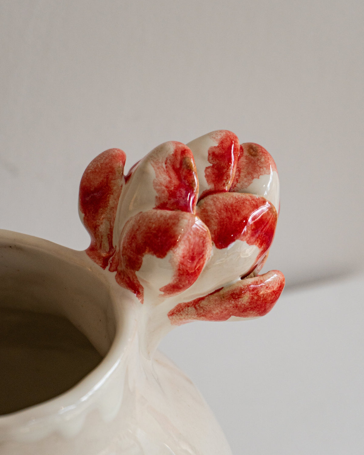A close-up of a glossy ceramic vessel with a sculptural red and white flower bud spout, set against a plain light gray background.