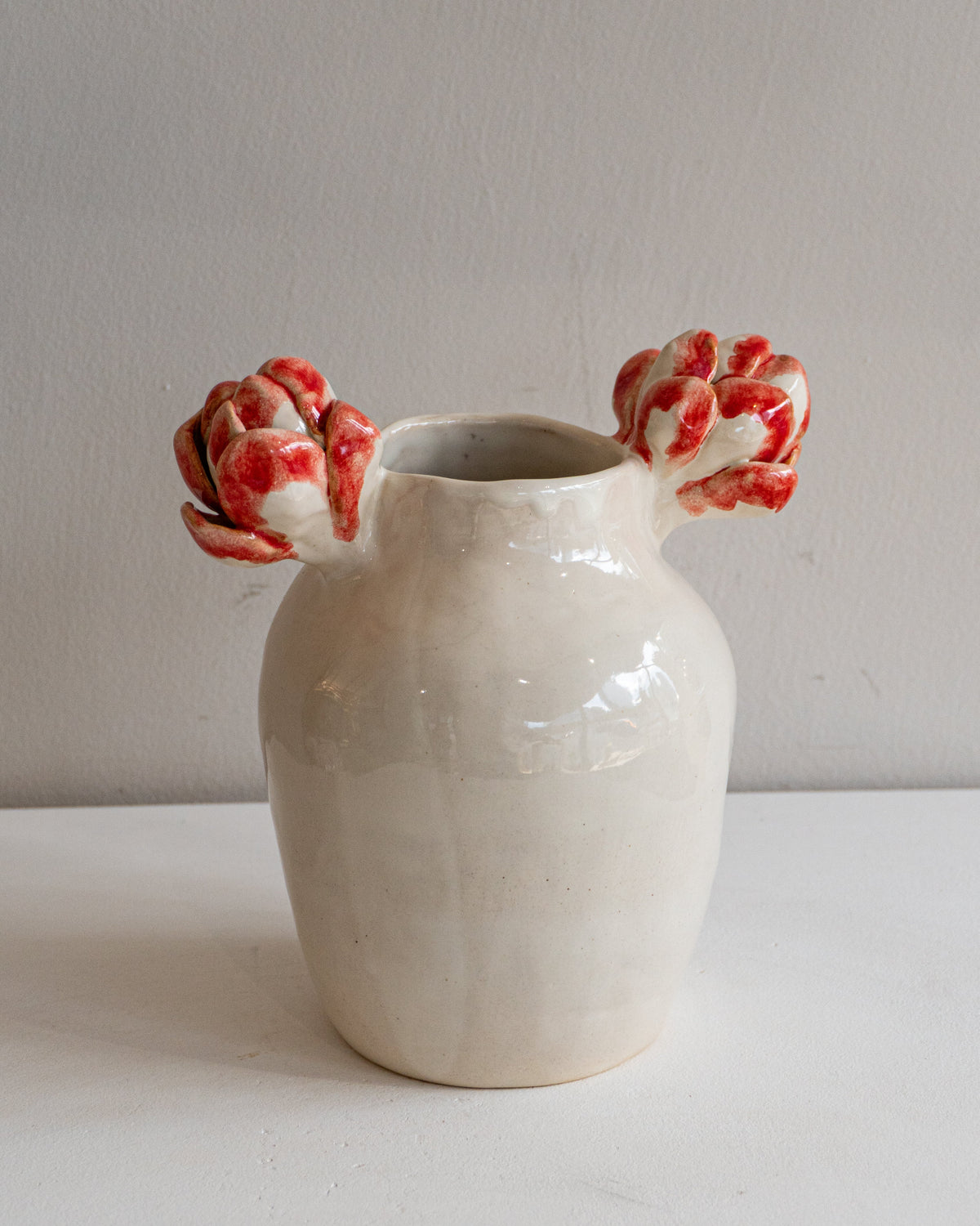 A handmade ceramic vase with a glossy off-white body and two red-and-white patterned tulip-shaped handles, set against a plain light gray background.