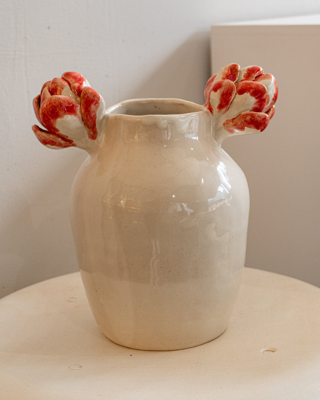 A handmade ceramic vase with a glossy cream-colored body and two sculptural red and white flower-like handles, displayed on a light wooden surface against a neutral background.