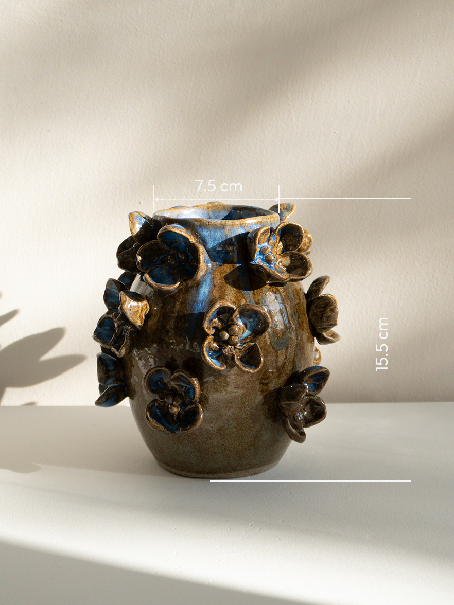 A small handmade ceramic vase with a brown and blue glaze, decorated with raised floral motifs, shown on a white surface against a beige wall with soft shadows. White measurement lines and text indicate dimensions.