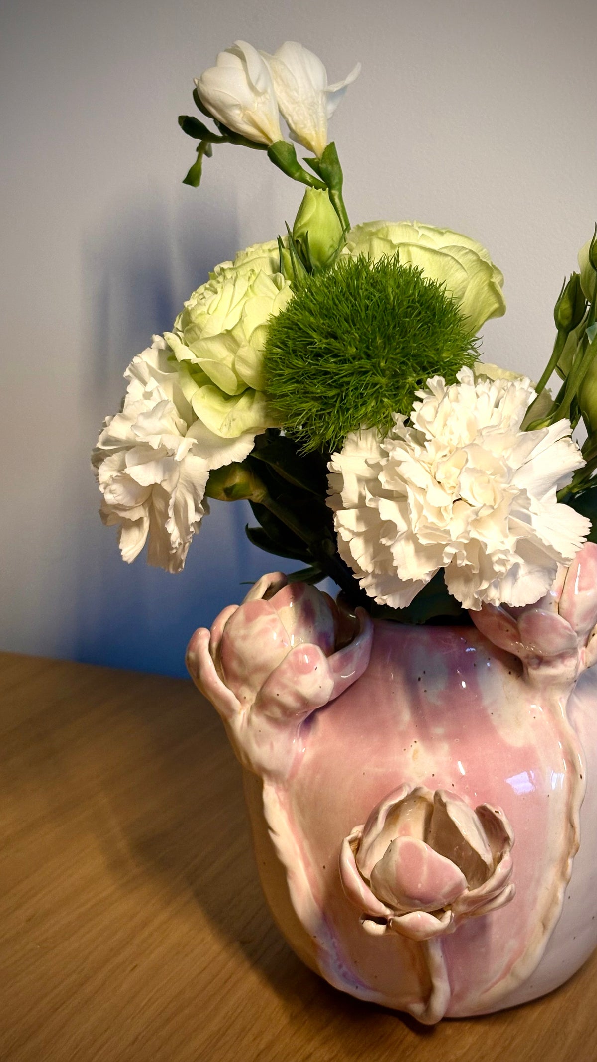 A pink ceramic flower vase with sculpted floral details, filled with white carnations, pale green lisianthus, and a green ditsy bloom, set on a wooden surface against a plain wall.